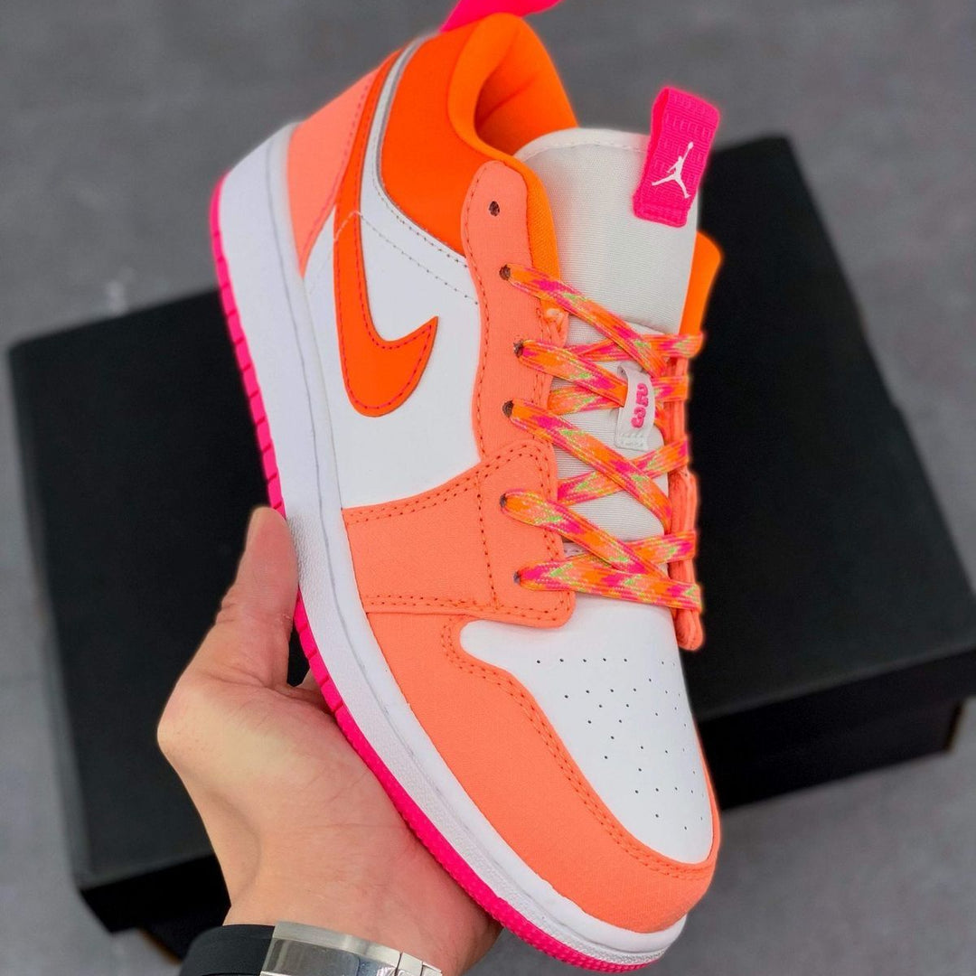 A hand holds a Nike Air Jordan 1 Retro Low sneaker in vibrant orange and white with hot pink accents, positioned in front of a black shoebox on a gray background.